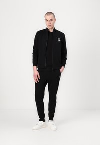 Black zip-up bomber jacket with a logo on the chest, paired with black joggers. Model wears white sneakers, standing against a plain background.