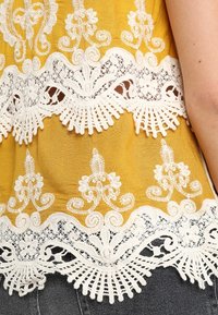 Sinappinkeltainen paita, jossa on monimutkaiset valkoiset pitsiyksityiskohdat helmassa, sisältäen kukka- ja aaltoilevia kuvioita teksturoidulla pinnalla.