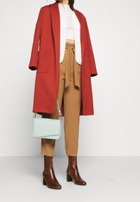 Röd, lång kappa med fickor, beige topp, bruna högmidjade byxor, bruna stövlar med krokodilskinnsmönster och en ljusblå handväska.