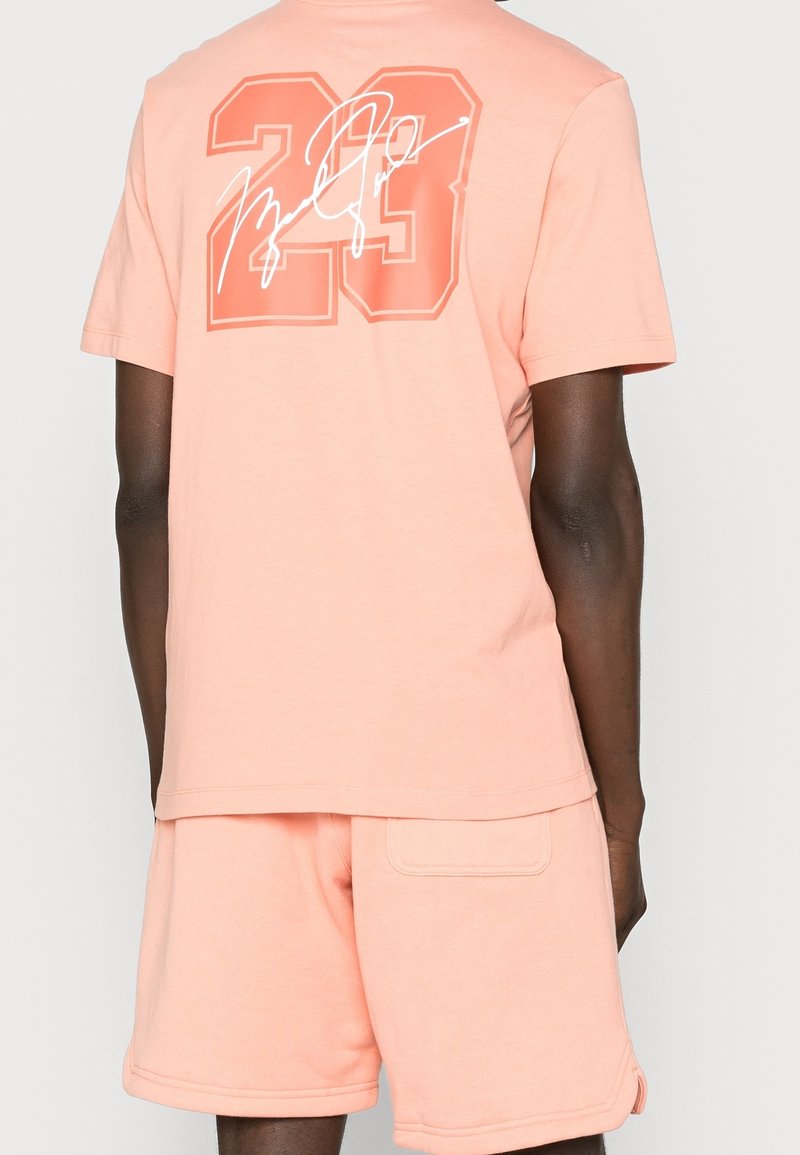 Camiseta de color melocotón con mangas cortas, que presenta un gran "23" y un gráfico de firma en blanco en la parte posterior. Textura de tela suave.