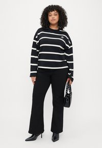 Sort og hvid stribet langærmet top med rund hals, parret med sorte flared bukser; holder en lille sort håndtaske med nitter på stropperne.