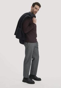 Dunkelburgunderfarbenes Sweatshirt, graue karierte Hose und schwarze Lederstiefel. Ein dunkler Mantel ist über die Schulter gelegt und zeigt die Schichten.