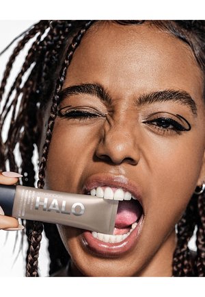 Hydratant teinté tout-en-un Smashbox Halo Healthy Glow dans un tube beige doux, tenu dans une bouche aux dents blanches avec un arrière-plan de cheveux tressés.