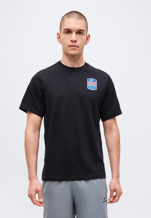 Homme portant un t-shirt noir Barcelona Nike et un short gris Nike, debout devant un fond clair uni.