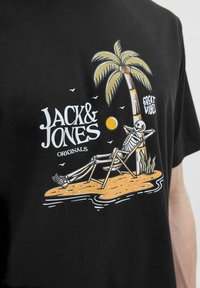 Schwarzes T-Shirt mit einer grafischen Illustration eines Skeletts, das entspannt in einem Liegestuhl unter einer Palme sitzt, mit dem Text "Jack & Jones Originals".