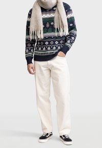 Mann in einem gemusterten navy-, grün- und beige-farbenen Pullover, beiger Schal, weißen Hosen und schwarzen Sneakers, der mit einer Hand in der Tasche steht.