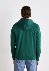 Felpa verde con cappuccio, realizzata in tessuto morbido. Presenta una vestibilità comoda e polsini a coste, abbinata a jeans di un blu chiaro.