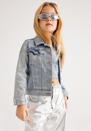 Chaqueta de mezclilla en azul claro con cierre de botones y bolsillos en el pecho, combinada con pantalones plateados brillantes y gafas de sol reflectantes.
