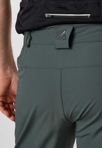 Gros plan d'une personne portant un pantalon vert foncé avec une étiquette de ceinture de marque et une chemise noire avec une petite poche zippée.