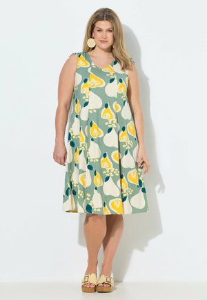 Kobieta ubrana w sukienkę bez rękawów z zielonym, żółtym i białym abstrakcyjnym wzorem, duże okrągłe kolczyki i beżowe sandały na otwartym palcu, stojąca na tle białej ściany.