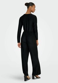 Mono negro con mangas largas, cintura elástica y diseño de pierna ancha. Fabricado con un material texturizado brillante. Combinado con tacones de bloque.