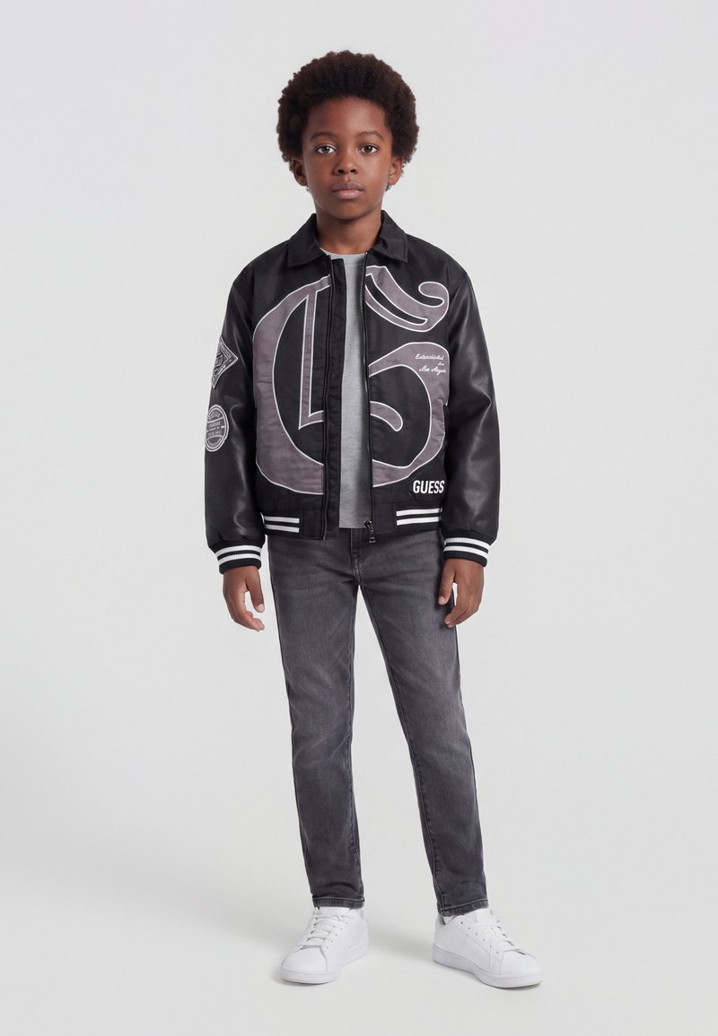 Garçon debout face à l’avant, portant une veste bomber noire et grise, un jean gris et des baskets blanches sur un fond uni clair.