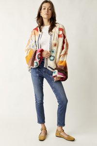 Γυναίκα που στέκεται με μπλε τζιν, λευκό πουκάμισο, πολύχρωμη χοντρή πλεκτή ζακέτα και ταμπά παπούτσια Mary Jane με ασημένια καρφιά, μπροστά σε απλό φόντο.