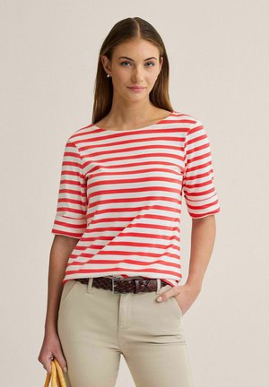 Kvinde iført en rød og hvid vandretstribet skjorte med albuelange ærmer, beige bukser og et flettet brunt bælte, der holder en gul taske.