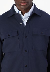 Marinblå skjortjacka med knappar, tillverkad av texturerat tyg, med två bröstfickor med svarta tryckknappar och en ljusblå skjorta med krage under.