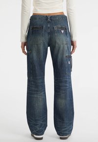 Person trägt lässige, weit geschnittene blaue Jeans mit mehreren Taschen und Reißverschlüssen, kombiniert mit einem weißen Langarmoberteil und weißen Schuhen.