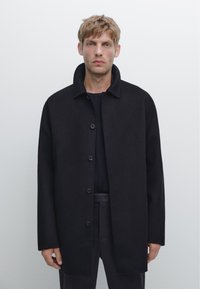 Cappotto di lana nera con colletto alto, chiusura frontale con bottoni e vestibilità Loose. Il tessuto ha una superficie liscia e un design pulito.