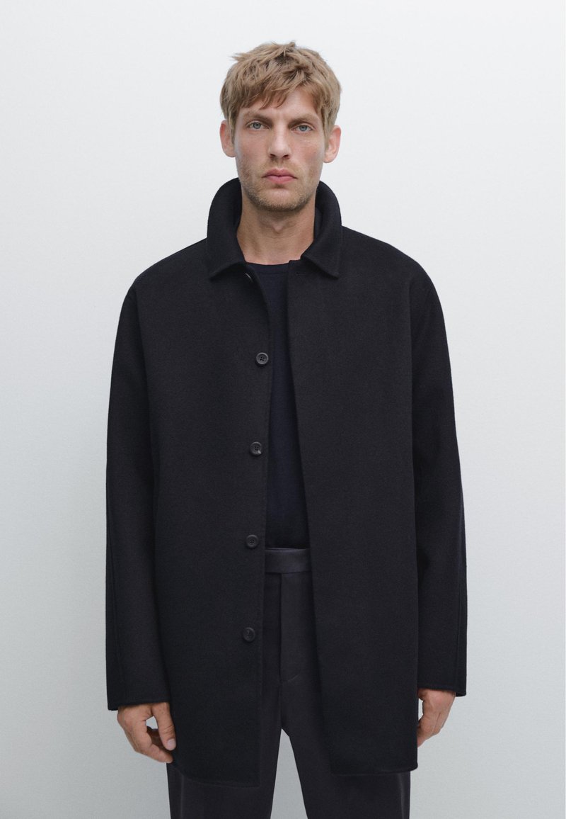 Cappotto di lana nera con colletto alto, chiusura frontale con bottoni e vestibilità Loose. Il tessuto ha una superficie liscia e un design pulito.