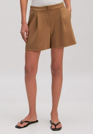 Vrouw draagt camelkleurige shorts met hoge taille en knoopsluiting, gecombineerd met zwarte platte sandalen, staand tegen een effen achtergrond.