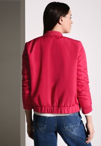 Veste bomber rose vif avec un col et des poignets côtelés, une taille élastiquée et une texture lisse, associée à un jean bleu foncé.