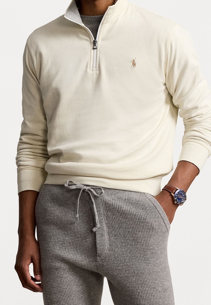 Sudadera crema con cremallera y puños acanalados, combinada con pantalones de chándal grises texturizados. Presenta un logotipo bordado en el pecho.