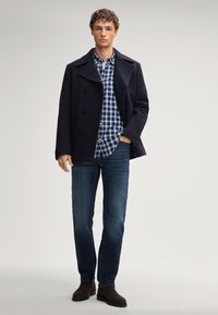 Cappotto navy sopra una camicia a scacchi blu e bianca, abbinato a jeans scuri e stivali di suede marrone. Il modello è in piedi contro uno sfondo semplice.