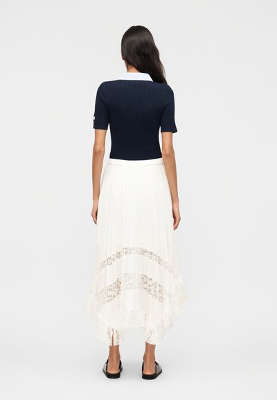Γυναίκα που φοράει μπλε σκούρη ριμπ κοντομάνικη μπλούζα με λευκό γιακά, λευκή πλισέ φούστα με δαντελένιες λεπτομέρειες και μαύρα παπούτσια χωρίς κορδόνια, γυρισμένη πλάτη.