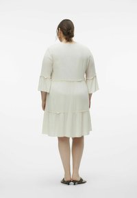Vestido beige claro con mangas acampanadas de tres cuartos, dobladillo de volantes en capas y una textura sutil. Llevado con sandalias negras, visto desde atrás.