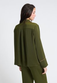 Oliwkowa bluzka z długimi rękawami o luźnym kroju, z plisą z tyłu, kontrastowym przeszyciem przy mankietach oraz gładką fakturą.