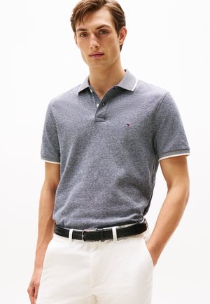 Junger Mann trägt ein graues kurzärmeliges Polohemd mit kleinem Logo und weiße Hose mit einem schwarzen geflochtenen Gürtel, steht mit einer Hand in der Hosentasche.