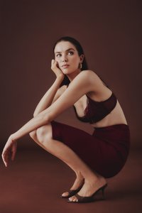 Vinröd spetsbehå och figurnära vinröd kjol med slät textur; modell hukande med öppna bruna högklackade mules, minimalistisk bakgrund.