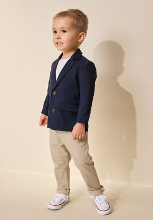 Jeune garçon debout, portant un blazer bleu marine, une chemise blanche, un pantalon beige et des baskets blanches sur un fond clair uni.