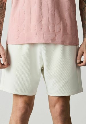 Mann trägt strukturierte weiße Shorts und ein rosa gemustertes Hemd, zeigt tätowierte Unterarme und Hände an den Seiten.