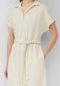 Abito camicia in lino beige con chiusura a bottoni, maniche corte arrotolate e cintura legata in vita. Presenta tasche laterali e una vestibilità comoda.