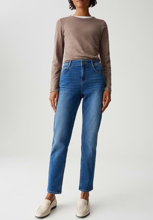Vrouw in een strakke blauwe jeans, taupe langemouwshirt over een witte top, en crèmekleurige instapschoenen, staande tegen een effen lichte achtergrond.