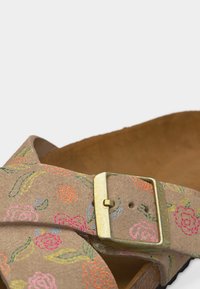 Κοντινή λήψη λουριού σανδαλιού από μπεζ σουέτ με πολύχρωμη κεντητική φλοράλ και μεγάλη τετράγωνη αγκράφα από ορείχαλκο.