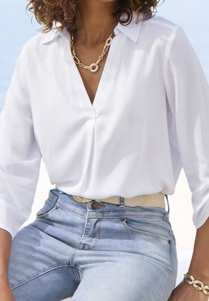 Chemisier blanc oversize à manches longues et décolleté en V, associé à un jean en denim bleu clair et une ceinture beige. Collier en chaîne dorée en accent.