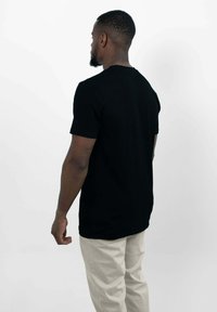 Zwarte katoenen t-shirt met korte mouwen en ronde hals; gedragen met lichtbeige broek; weergegeven van achteren tegen een witte achtergrond.