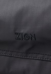 Black textured fabric featuring the embroidered brand name "ZIGN" in a contrasting black thread, with visible stitching details.