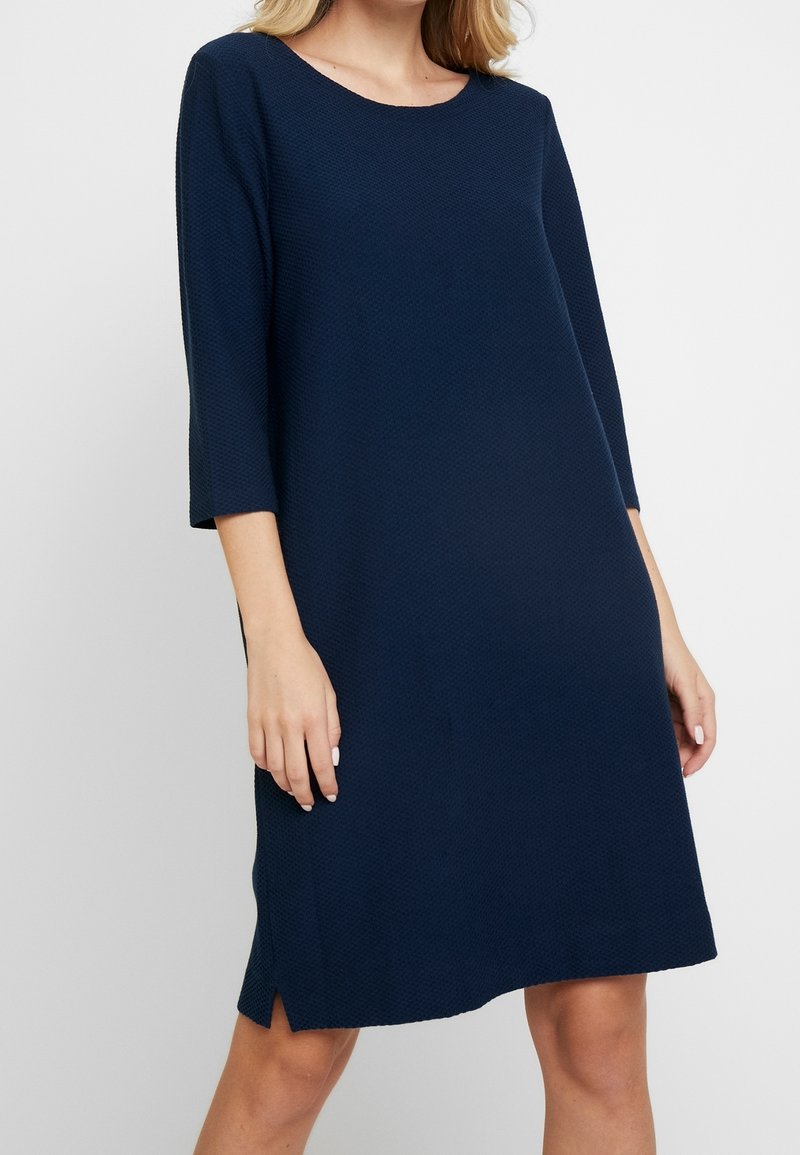 Robe bleu marine avec un motif texturé, encolure ronde, manches trois-quarts et fentes sur les côtés. Silhouette décontractée en A.