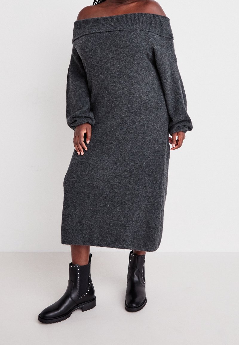 Personne portant une robe-pull longue gris foncé épaules dénudées et des bottines en cuir noir cloutées, debout devant un fond uni clair.