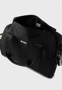 Μαύρη νάιλον τσάντα duffel με διπλές υφασμάτινες λαβές, κλείσιμο με φερμουάρ και ορατό λογότυπο. Διαθέτει ευρύχωρο εσωτερικό και ελαστική πλαϊνή τσέπη.