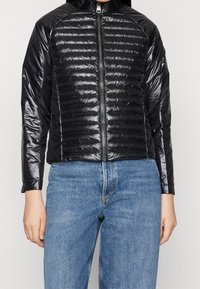 Svart puffjacka med en glänsande yta, med horisontellt kviltade sektioner, en framknäppning och långa ärmar, som är ihopparad med blå denimjeans.