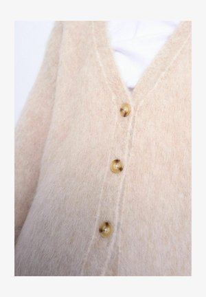Zachte beige, pluizige cardigan met drie ronde, lichtbruine knopen dichtgemaakt over een wit shirt.