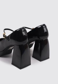 Zapatos de charol negros con tacón cuadrado. Cuentan con una delgada correa para el tobillo y detalles en hebilla dorada. Textura suave y brillante.