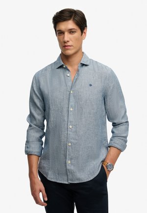 Mężczyzna o ciemnych włosach, ubrany w jasnoniebieską, długorękawową koszulę na guziki z podwiniętymi rękawami, ciemne spodnie i zegarek na lewym nadgarstku.