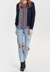 Marinblå cardigan över en mönstrad blus med blå, rosa och vita ränder; slitna ljusblå jeans med knäskärningar; svarta ankelstövlar.