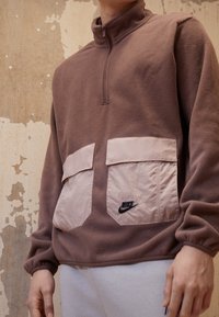 Pull en polaire marron avec un col haut, doté de deux poches avant rose clair et d'un logo Nike noir. Texture lisse et coupe décontractée.