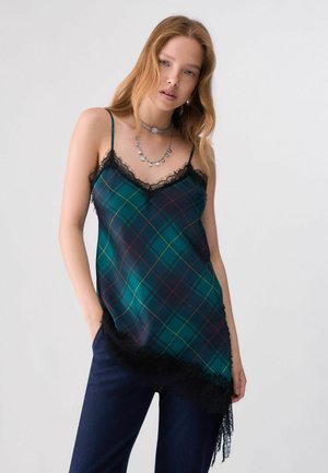 Femme aux longs cheveux portant un débardeur à carreaux verts et bleus avec une bordure en dentelle noire, un pantalon sombre et des colliers en argent superposés, face à l'avant.
