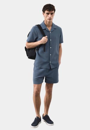 Junger Mann trägt ein blaues Hemd mit kurzen Ärmeln und passenden Shorts, schwarzen Rucksack über einer Schulter und marineblaue Turnschuhe.